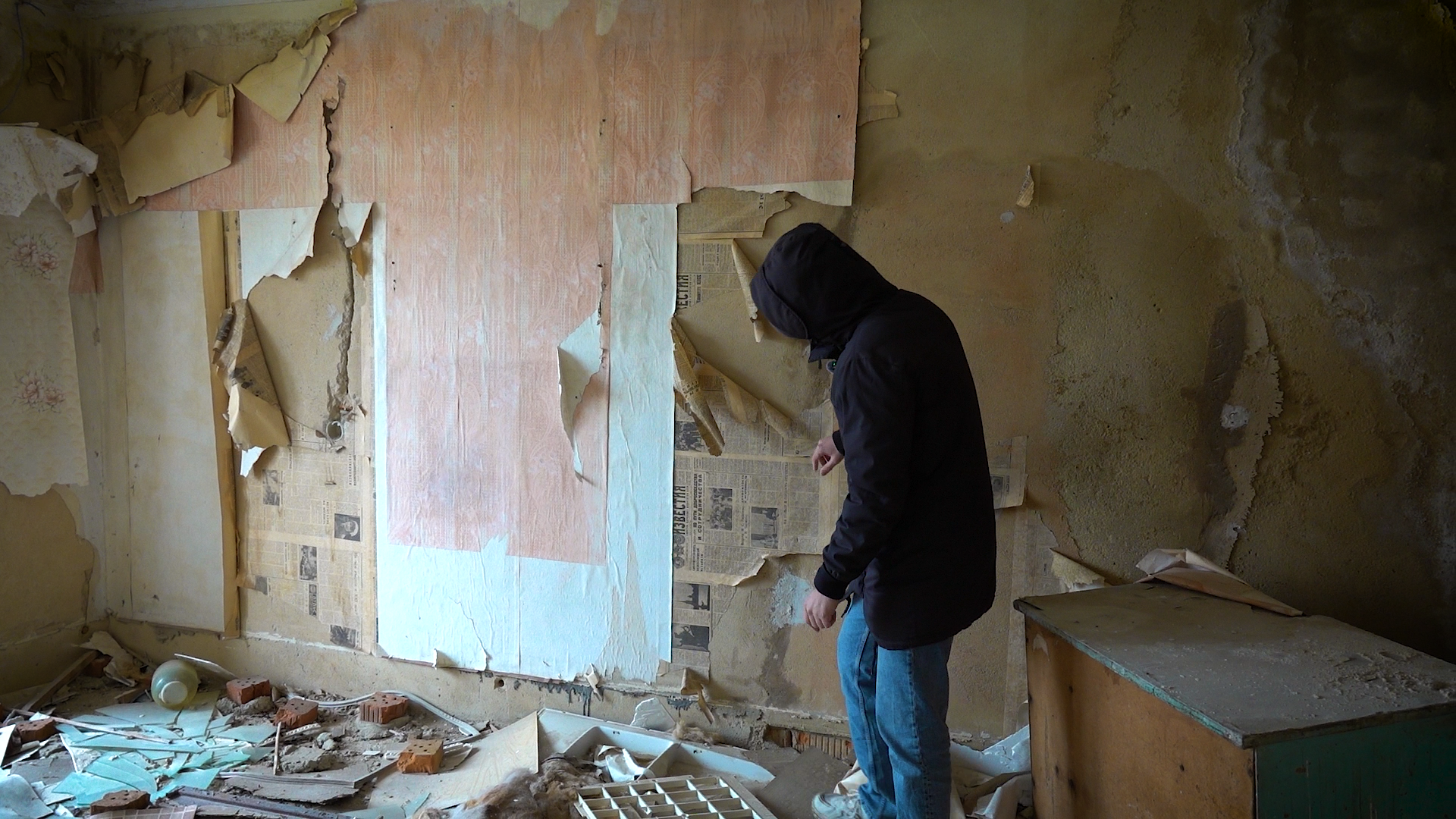 Журналист в комнате брошенной в Красногорском пятиэтажке, на стенах ободранные обои, под ними видны газеты